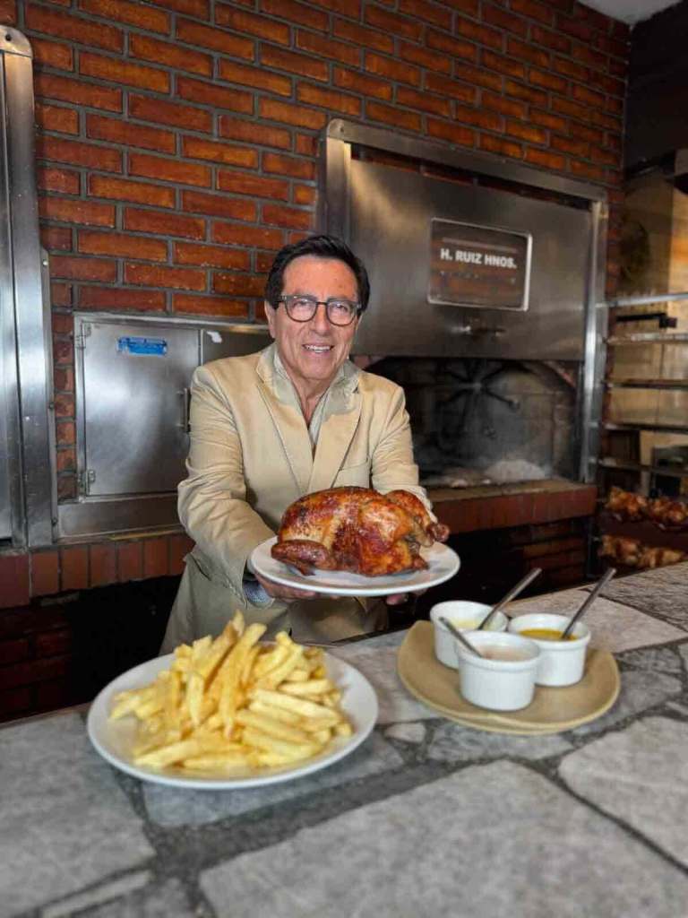 Un hombre sonriente sostiene un plato con pollo a la brasa, acompañado de papas fritas y salsas, en un ambiente de restaurante con paredes de ladrillo.