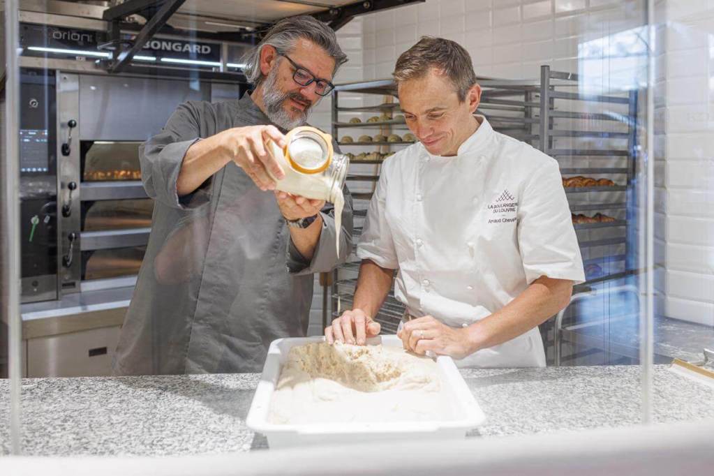 Dos panaderos trabajando en la panadería del Museo del Louvre, uno vertiendo masa madre Vitus en un recipiente mientras el otro amasa la mezcla.
