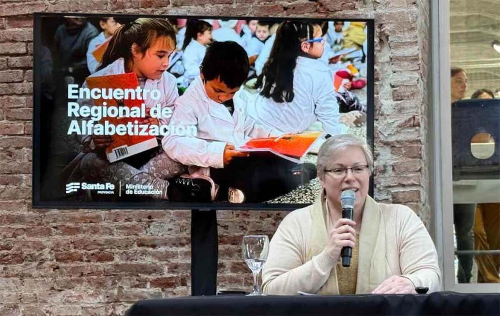Una presentadora habla en el Encuentro Regional de Alfabetización, con una pantalla detrás que muestra a niños participando en actividades de lectura.
