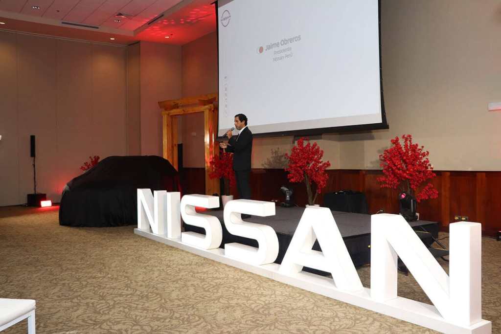 Presentación de la Nueva Nissan Qashqai 2025 en un evento, con un presentador hablando frente a un grupo, un vehículo cubierto en el fondo y decoración de flores rojas.