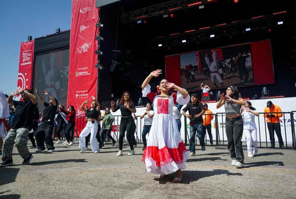 Grupo de personas bailando en un escenario al aire libre durante la feria Perú Mucho Gusto Tacna 2025. La imagen muestra a una mujer vestida con un traje tradicional blanco y rojo, con otros asistentes participando en una danza colectiva.