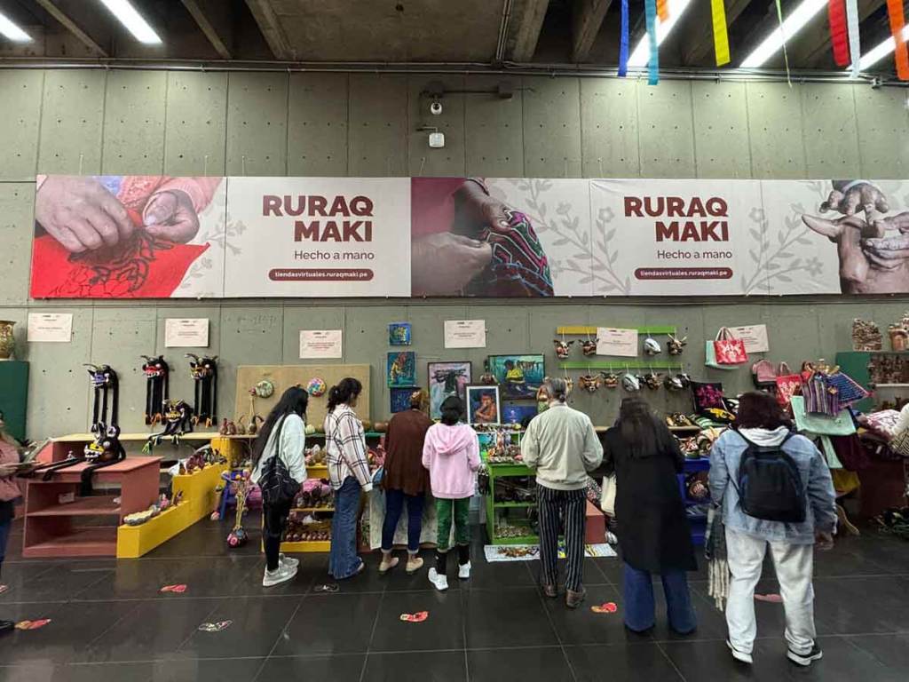 Un grupo de mujeres artesanas se encuentra en la feria Ruraq Maki, observando y mostrando diversas artesanías hechas a mano, con un enfoque en la herencia cultural y la sostenibilidad.