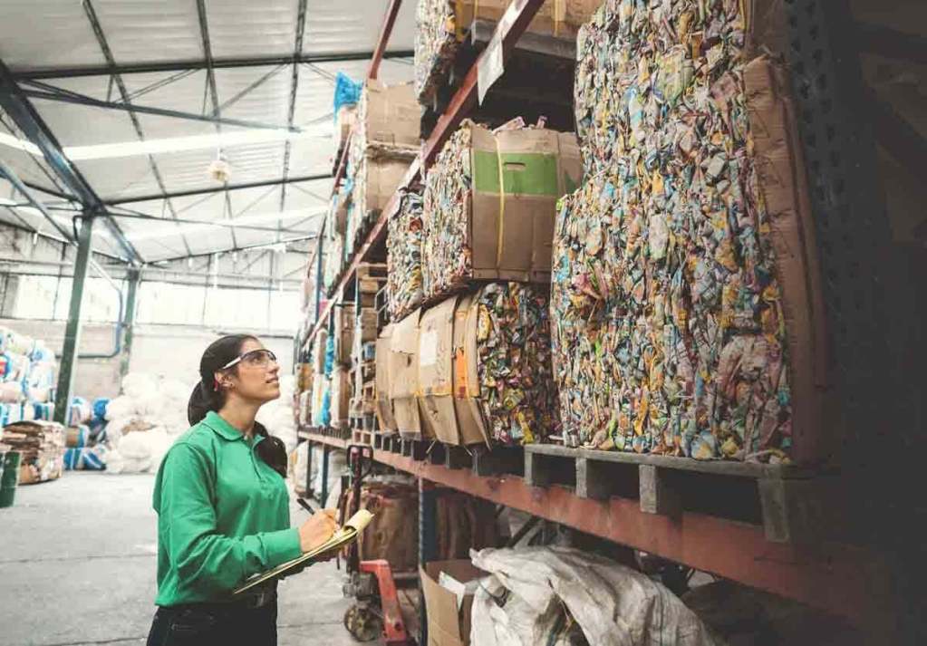 Una mujer con una camisa verde y gafas de seguridad revisa pilas de cartones reciclados en un almacén.