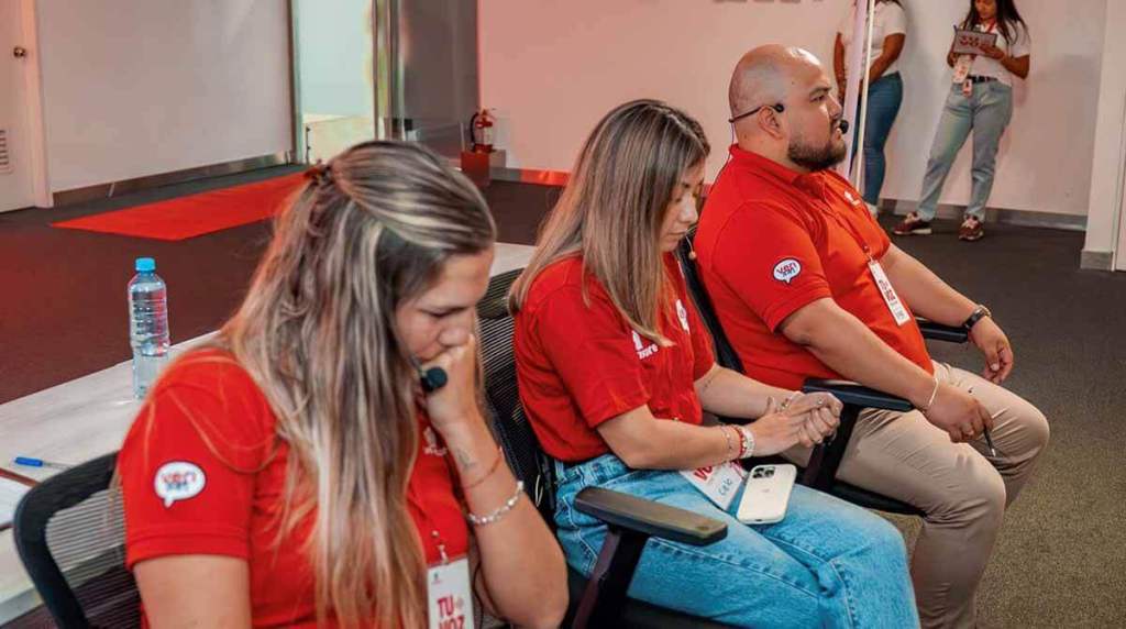 Tres evaluadores en una audición de reclutamiento, enfocados en una simulación de atención al cliente, sentados en sillas con camisetas rojas, en un ambiente profesional.