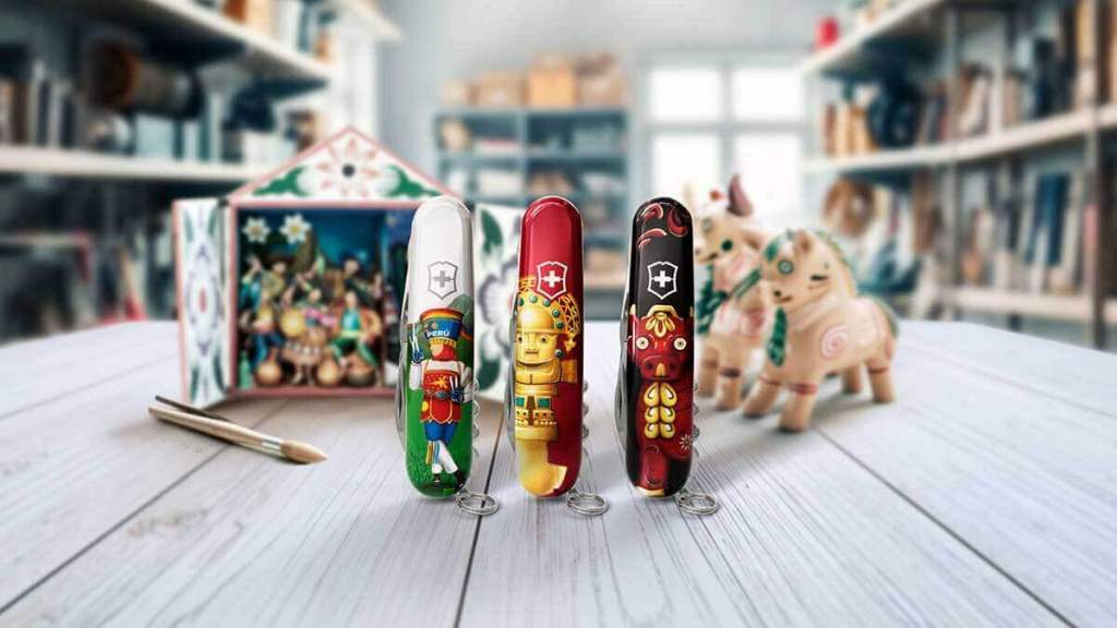 Tres navajas suizas de edición exclusiva de Victorinox, decoradas con símbolos de la cultura peruana, sobre una mesa de madera. Al fondo, se observa un retablo y dos figuras de artesanía peruana.