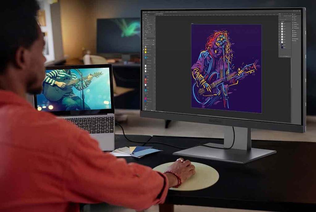 Una persona trabajando en una computadora mientras edita una ilustración en un monitor de 27 pulgadas, con una laptop mostrando un video musical.