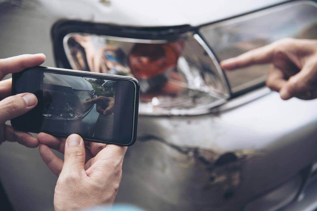 Una persona tomando una foto con un teléfono móvil de un auto con daños en el faro mientras otra persona señala el daño.