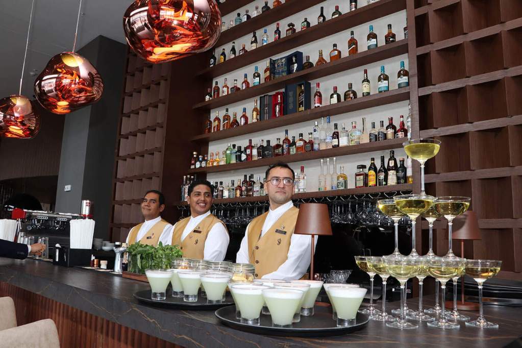 Tres bartenders en el bar del hotel Wyndham Grand Costa del Sol Lima Airport, con botellas de bebidas alcohólicas en estantería detrás y copas en la barra.