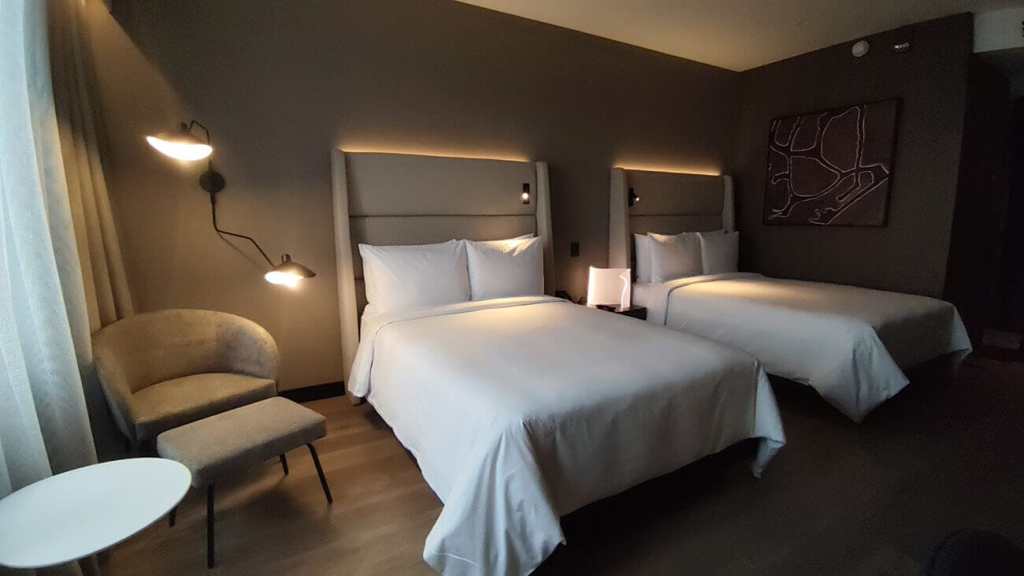 Habitación doble del hotel Wyndham Grand Costa del Sol Lima Airport con dos camas grandes, un sillón y mesa pequeña en una atmósfera moderna y elegante.