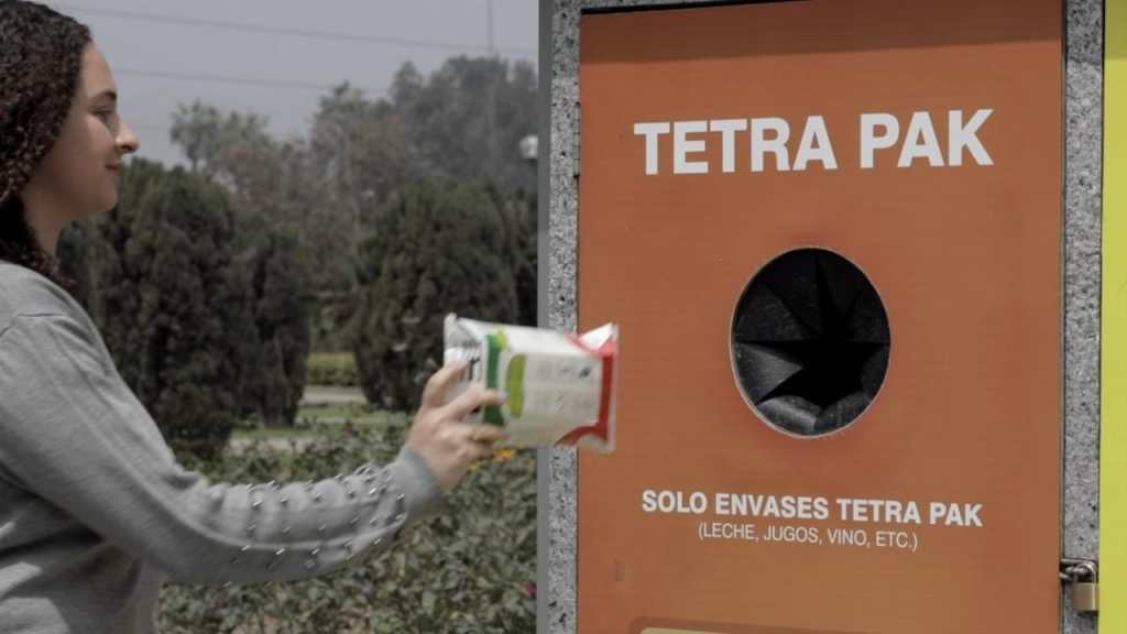 Una persona depositando un envase de Tetra Pak en un contenedor de reciclaje, con información visible sobre el reciclaje de envases Tetra Pak.
