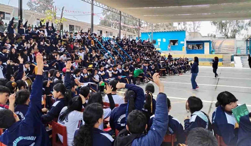Un grupo numeroso de estudiantes levantando las manos durante una actividad en un espacio escolar al aire libre, con docentes al frente interactuando.