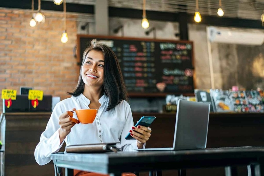 Mujer sonriente sosteniendo una taza de café en una cafetería, mirando su teléfono móvil, con una laptop en la mesa.