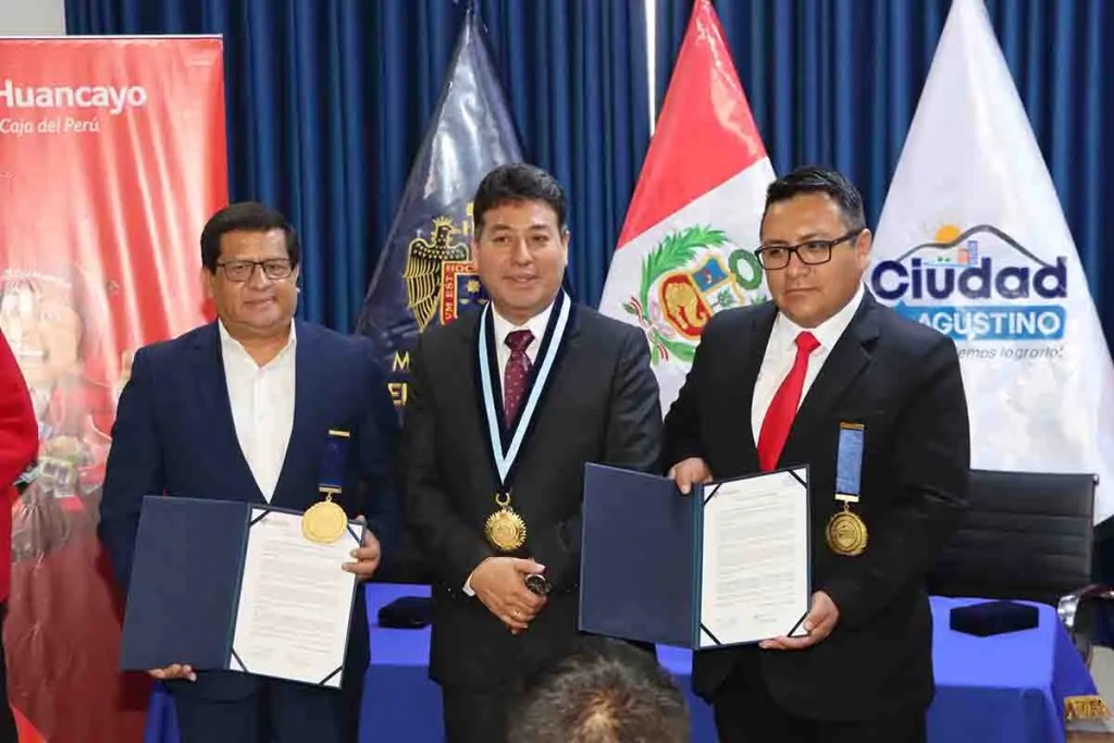 Tres hombres en una ceremonia formal, sosteniendo documentos, con banderas peruanas y logos visibles de la Municipalidad y Caja Huancayo de fondo.