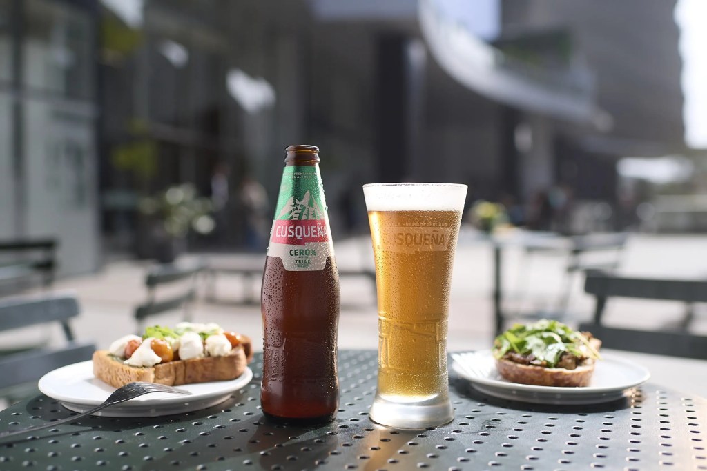 Una botella de Cerveza Cusqueña Trigo Cero Alcohol junto a un vaso de cerveza y platos con comida en una mesa al aire libre.