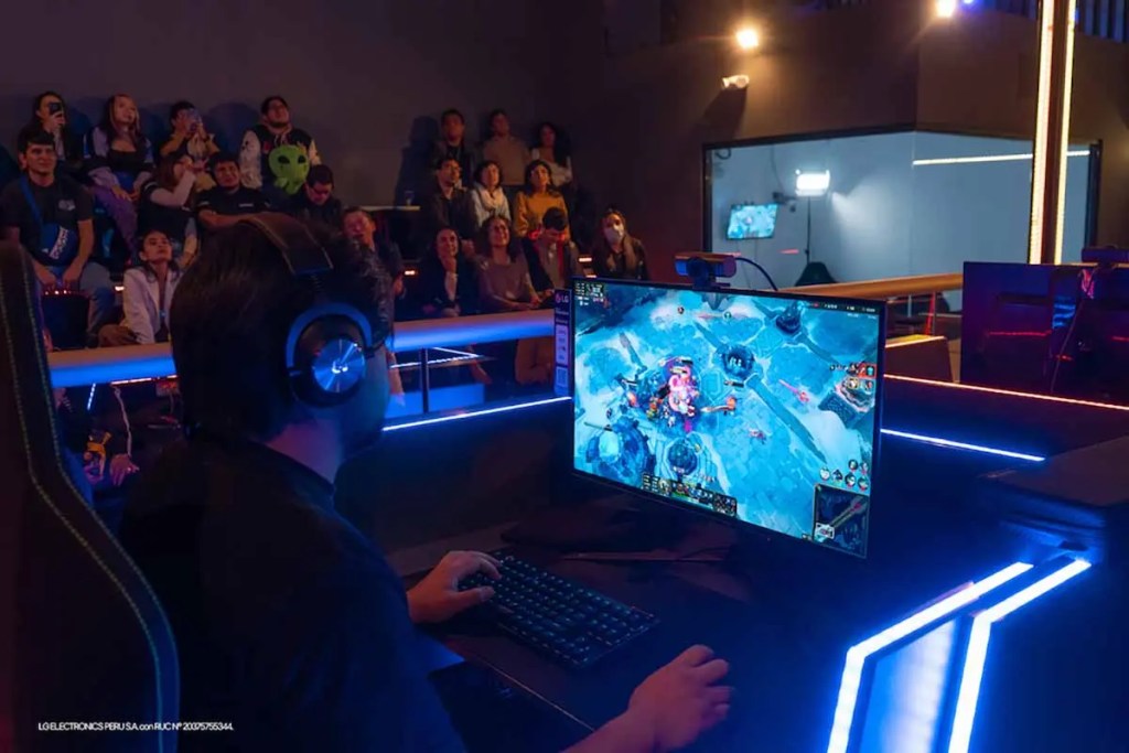 Un jugador concentrado frente a un monitor, participando en un torneo de videojuegos, mientras una audiencia lo observa desde diferentes asientos.