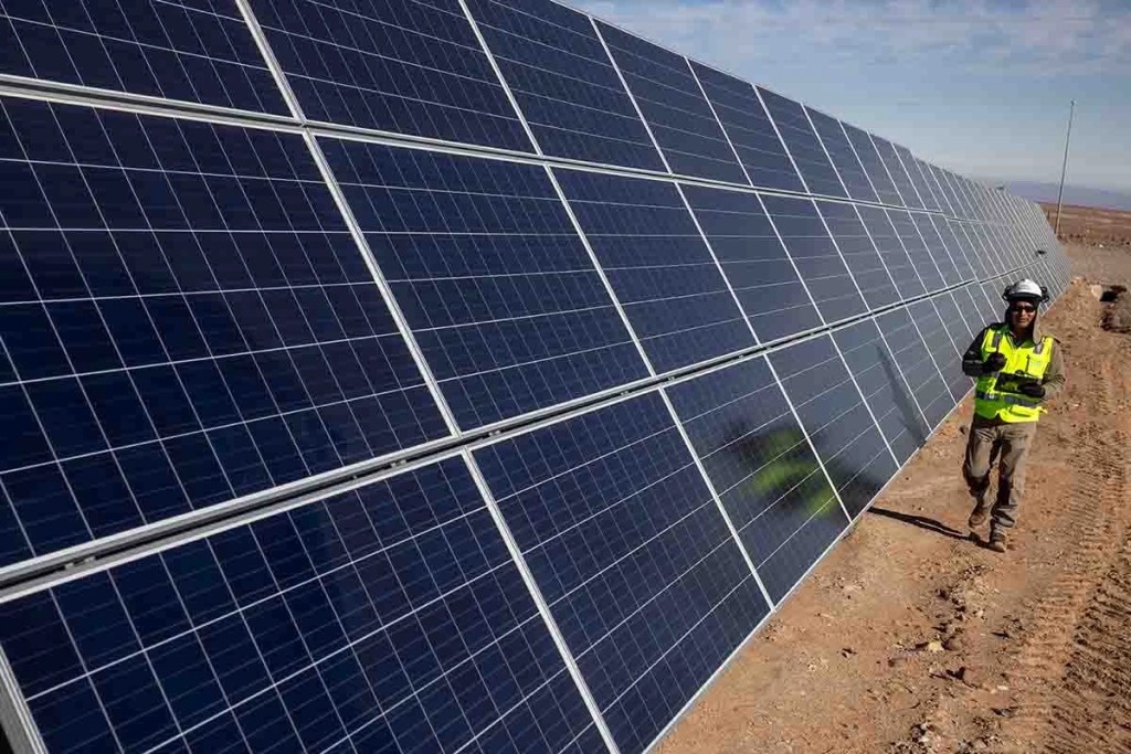 Un trabajador en un sitio de construcción de paneles solares, con una fila de paneles solares en el fondo, bajo un cielo despejado.