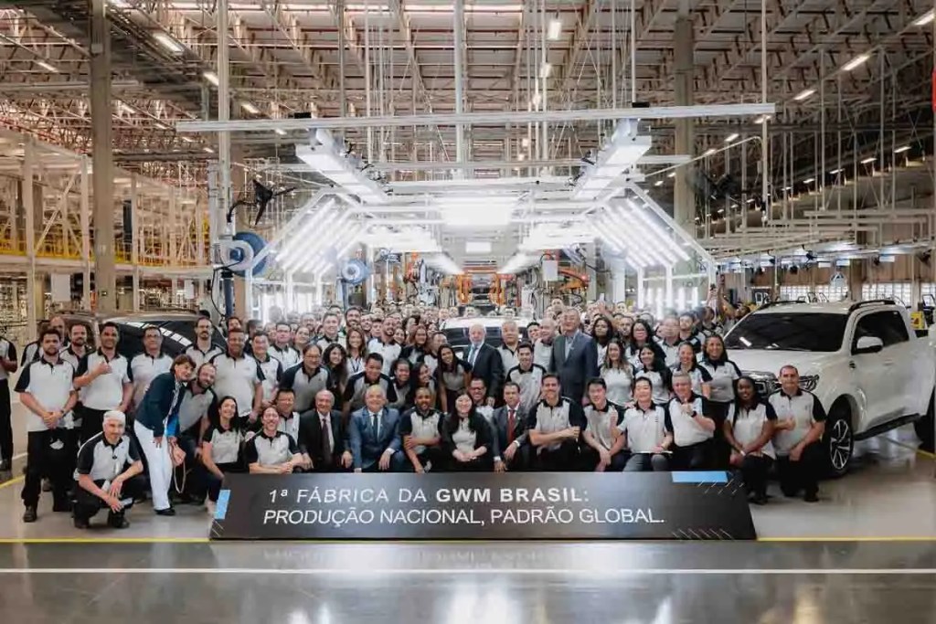 Celebración de apertura de la primera planta de GWM en Brasil, con un grupo de empleados posando frente a un vehículo en un entorno de fabricación moderna.