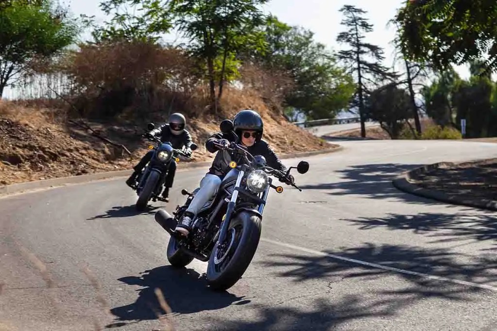 Dos motocicletas en una curva de carretera, una de ellas con un conductor y la otra con un pasajero, rodeadas de vegetación y un paisaje natural.