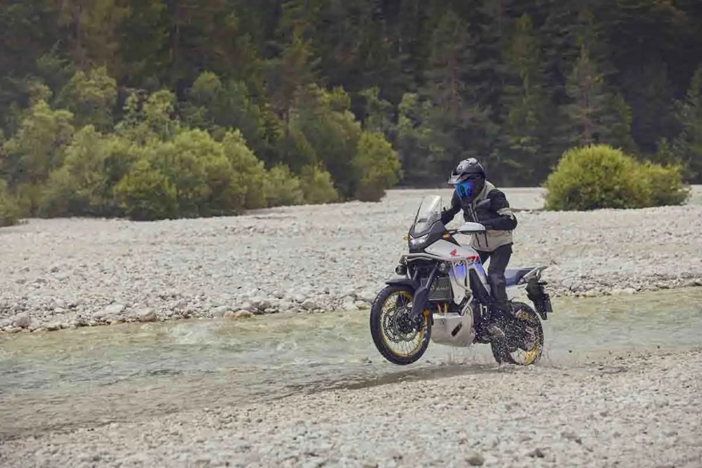 Motocicleta Honda XL750 Transalp cruzando un arroyo en un entorno natural con vegetación densa.