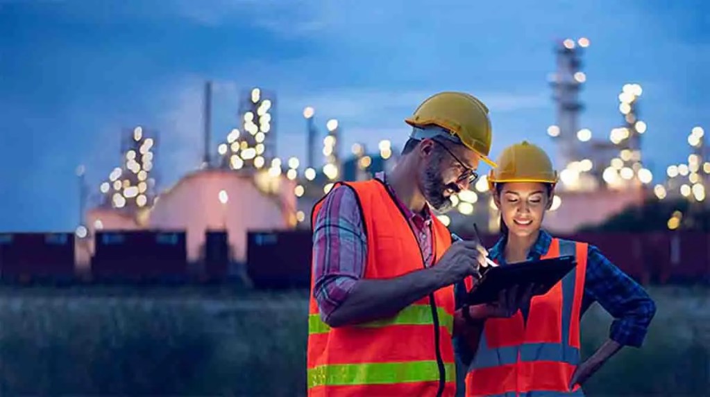 Dos trabajadores en un entorno industrial, revisando información en una tablet durante la noche, con luces brillantes de una planta en el fondo.