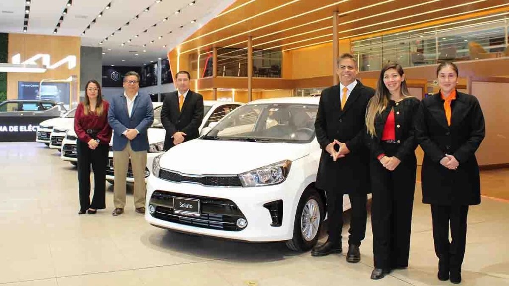 Grupo de seis personas posando junto a un vehículo Kia Soluto AT LX Plus en un concesionario, con varios modelos de Kia visibles al fondo.