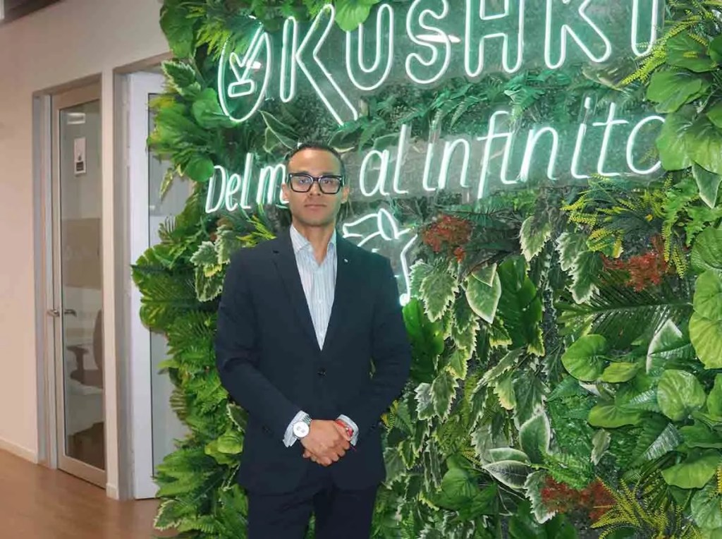 Persona de pie frente a un muro decorado con plantas y el logo de Kushki iluminado, que representa una plataforma de pagos en Perú.