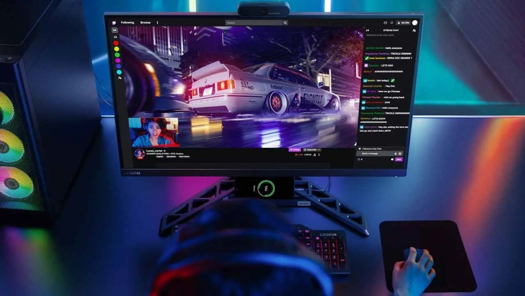 Vista de un monitor gaming mostrando una transmisión en vivo con un jugador interactuando, acompañado de un chat en la pantalla. Se observa un teclado iluminado y un mouse, todo en un ambiente de juego moderno.