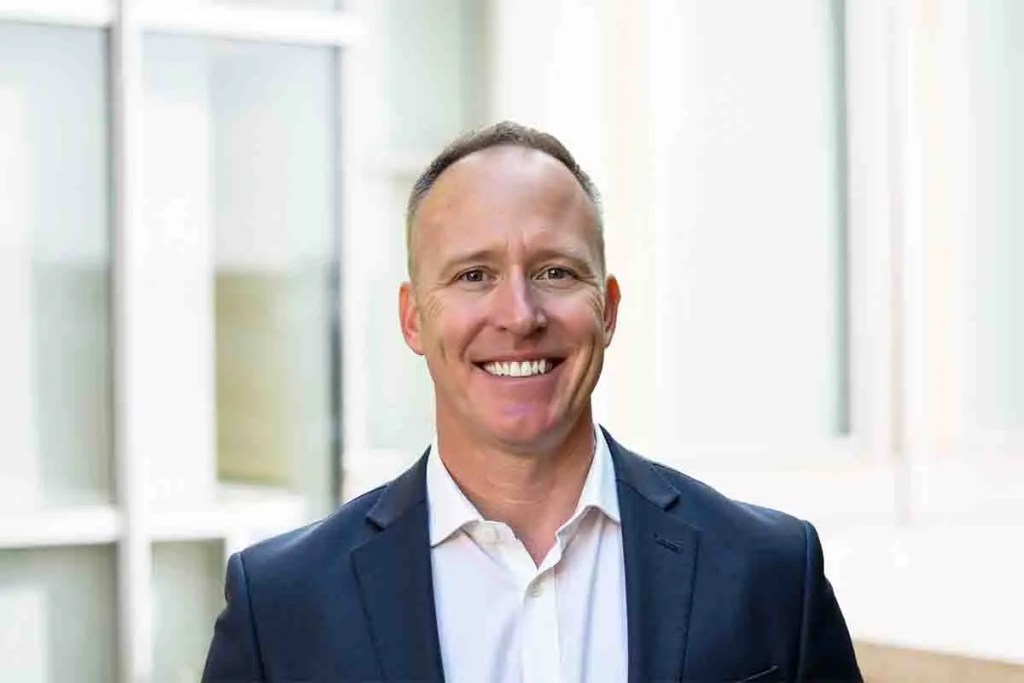 Retrato de Matthew Brown, nuevo Director Financiero de Tenable, sonriendo y vistiendo un traje oscuro frente a un fondo de ventana iluminada.