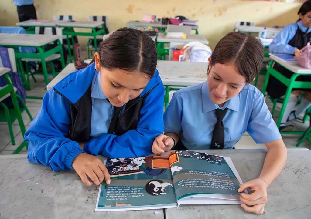 Dos escolares revisando un libro en un aula, con escritorios y sillas verdes. Una de las estudiantes lleva un uniforme azul con una chaqueta negra, mientras que la otra viste una camisa azul con corbata negra.