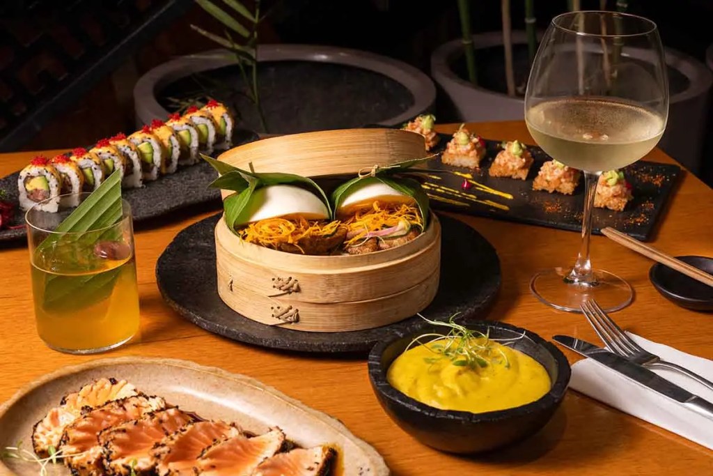 Una mesa con una variedad de platos asiáticos, incluyendo un bao en una vaporera, sashimi, una bebida amarilla, y un plato de sushi. El ambiente es elegante y acogedor, ideal para disfrutar de la cocina fusión peruano-asiática.