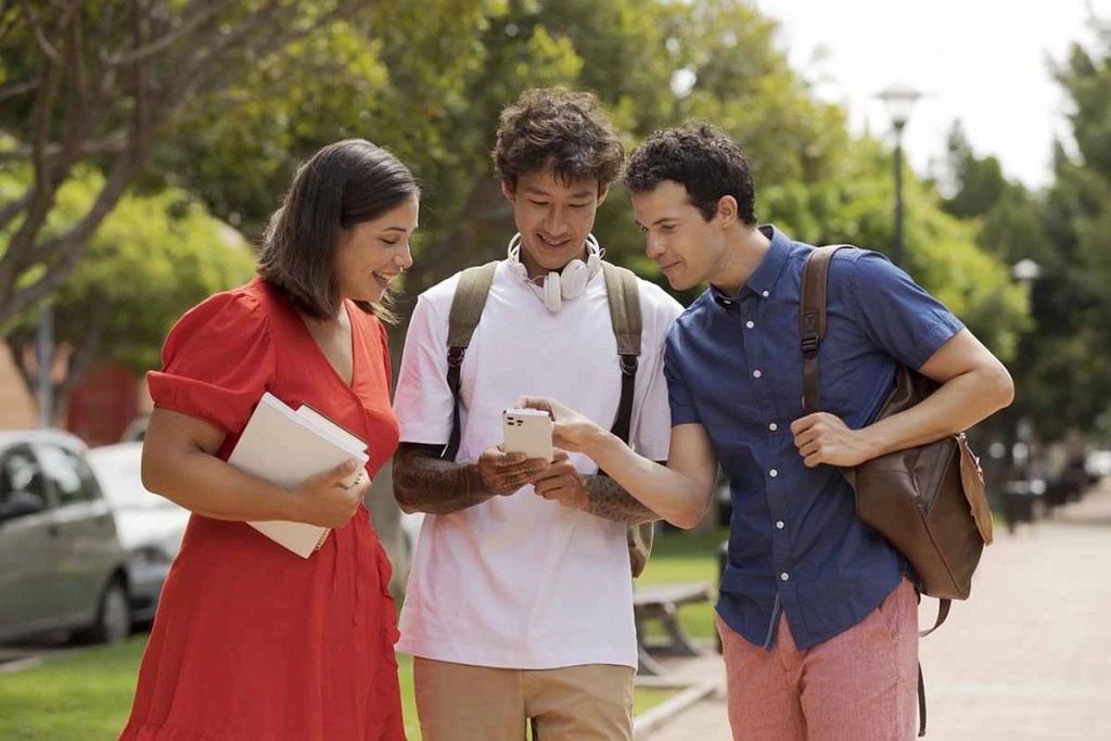 Tres estudiantes se agrupan al aire libre, mirando una pantalla de smartphone y sonriendo. Uno sostiene libros y los otros tienen mochilas, mostrando un ambiente universitario moderno.
