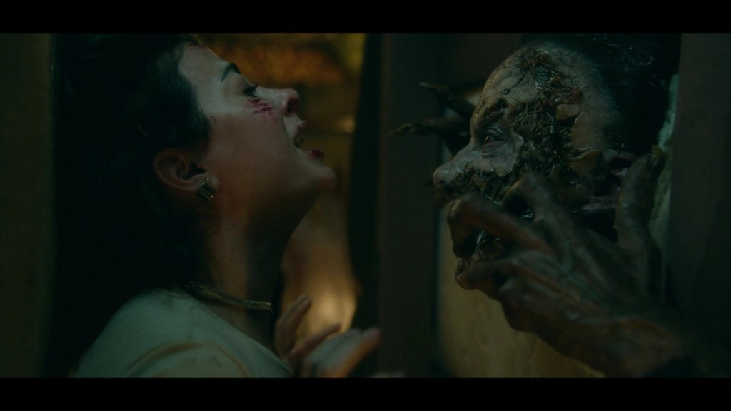 Una mujer con heridas en el rostro grita mientras enfrenta una criatura aterradora con una apariencia grotesca, en un ambiente oscuro y tenso que evoca un horror psicológico.