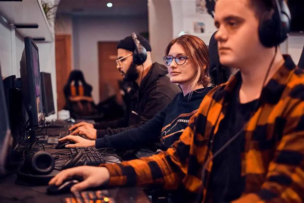 Tres personas concentradas jugando videojuegos en computadoras, equipadas con auriculares, en un entorno moderno y bien iluminado.