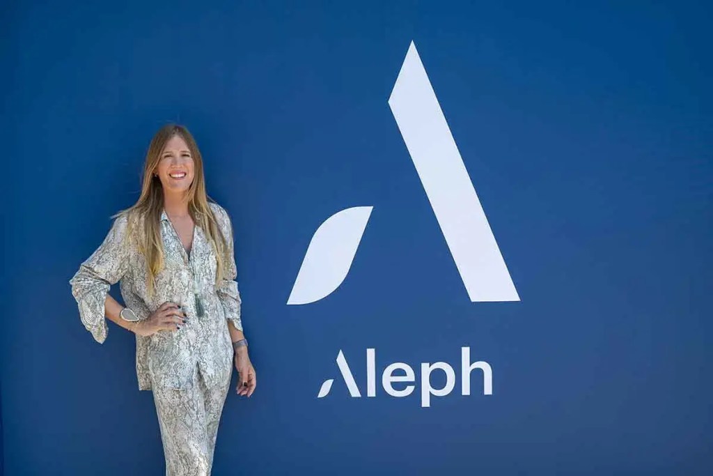 Mujer de pie junto a un logo de Aleph en una pared azul, sonriendo y luciendo una camisa de estilo elegante.