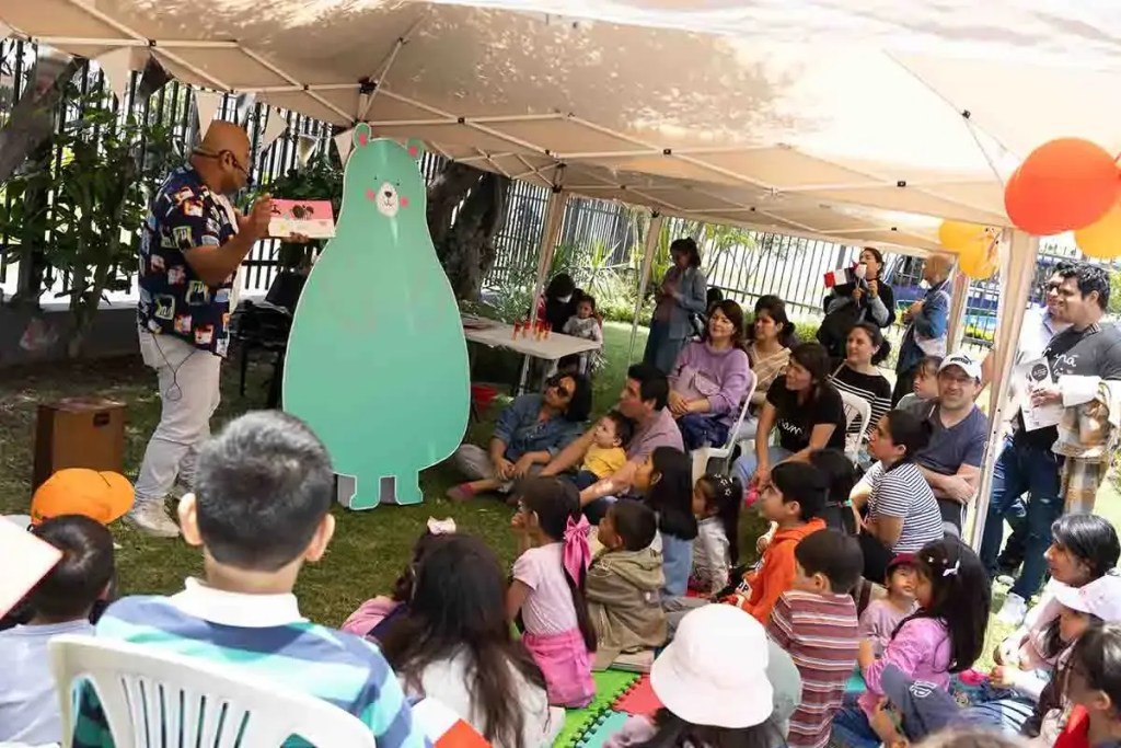 Un narrador sostiene un libro frente a un grupo de niños y adultos sentados en sillas y en el suelo, mientras un gran oso de cartón está presente en el escenario al aire libre bajo una carpa.