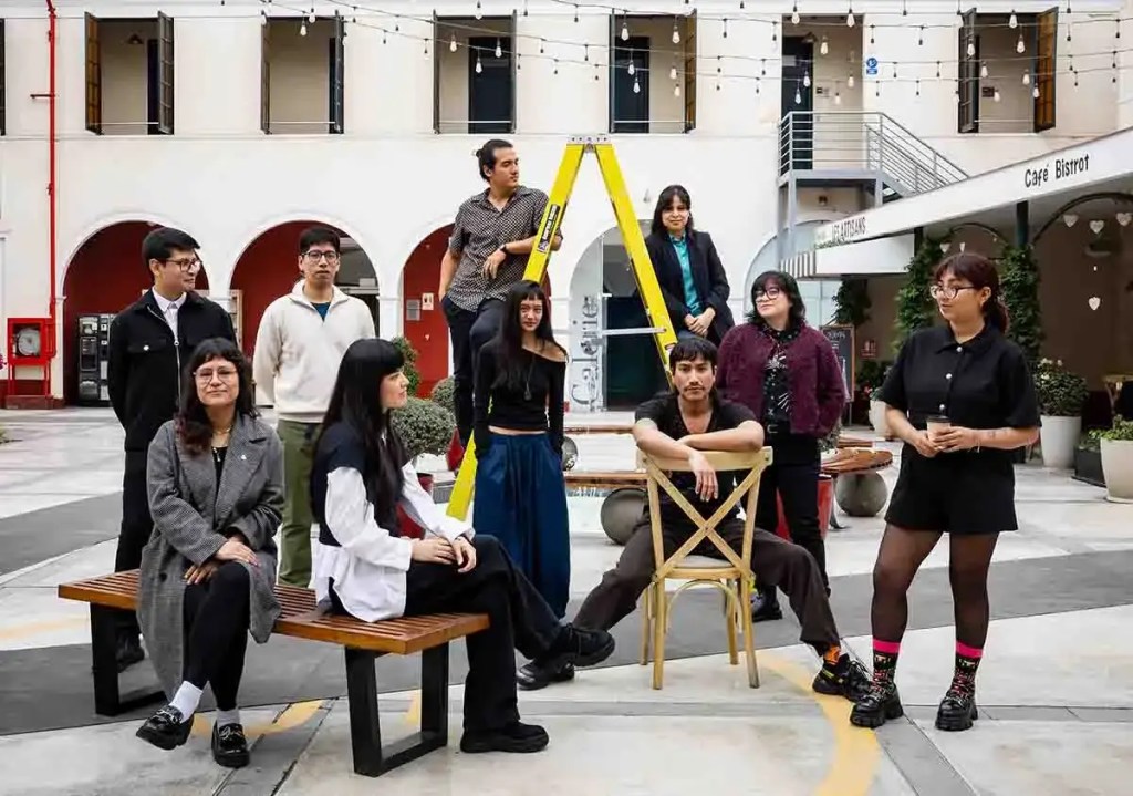 Diez jóvenes artistas peruanos posan juntos en un espacio al aire libre, algunos sentados y otros de pie, con una escalera amarilla en el fondo.