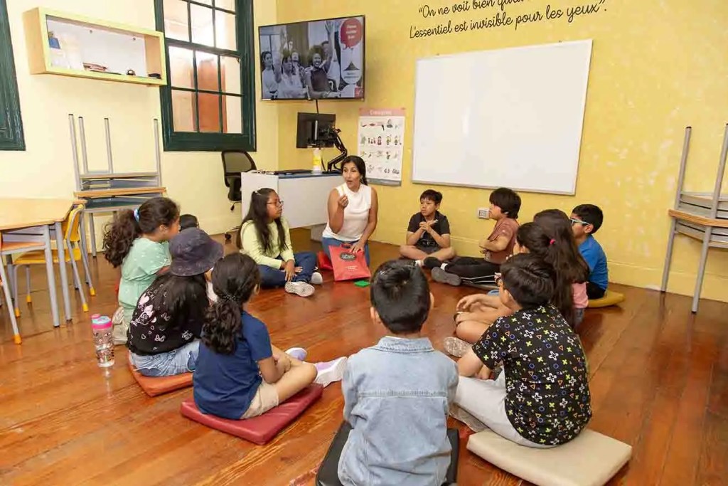 Un grupo de niños sentados en un salón de clases mientras una mujer les habla, con una pantalla y una pizarra blanca en el fondo.