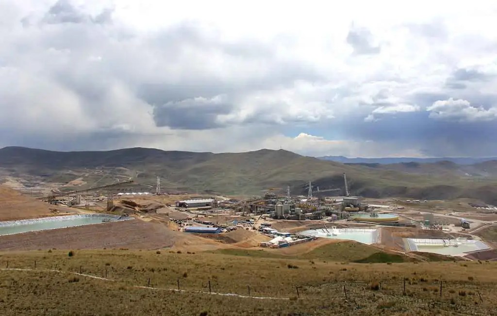 Panorama de un paisaje minero con instalaciones industriales y estanques en el suelo, en un entorno montañoso bajo un cielo nublado.