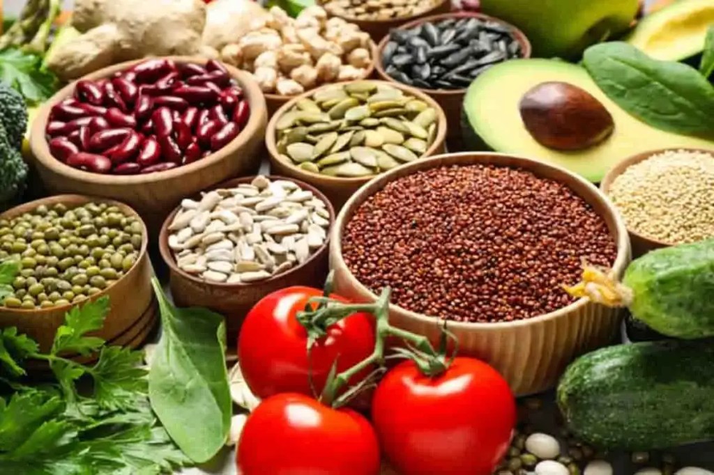 Variedad de alimentos sobre una mesa, incluyendo legumbres, semillas, verduras y frutas en recipientes y sueltos.