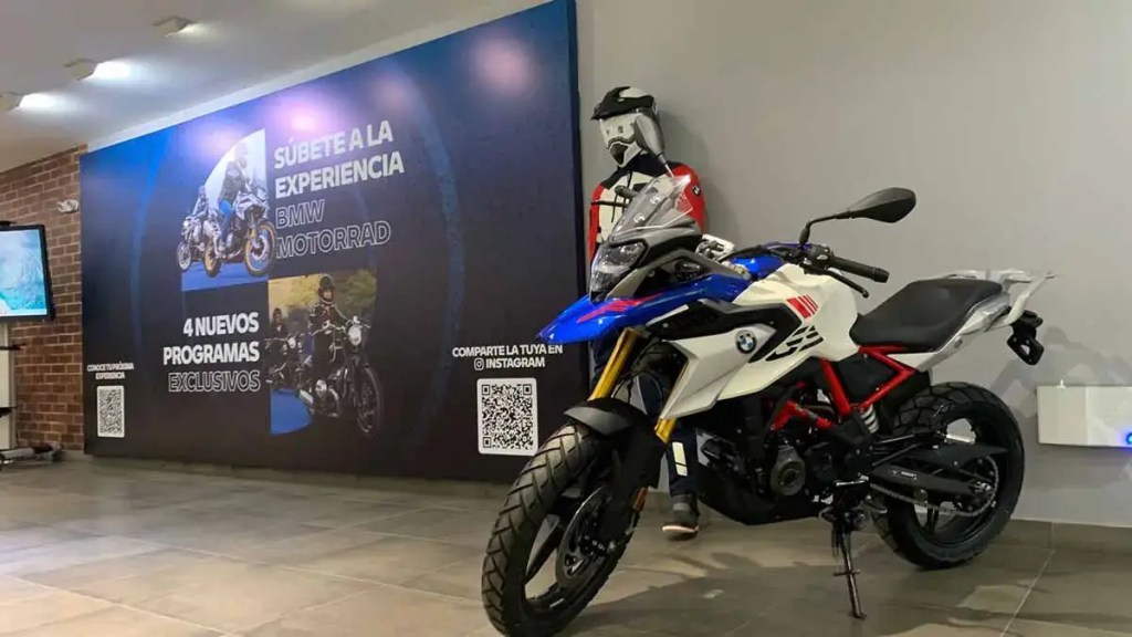 Imagen de una motocicleta BMW con un diseño deportivo y moderno, situada en un ambiente con un fondo que promociona el programa "Súbete a la Experiencia BMW Motorrad". Se observa un maniquí vestido con ropa de motociclista al fondo.