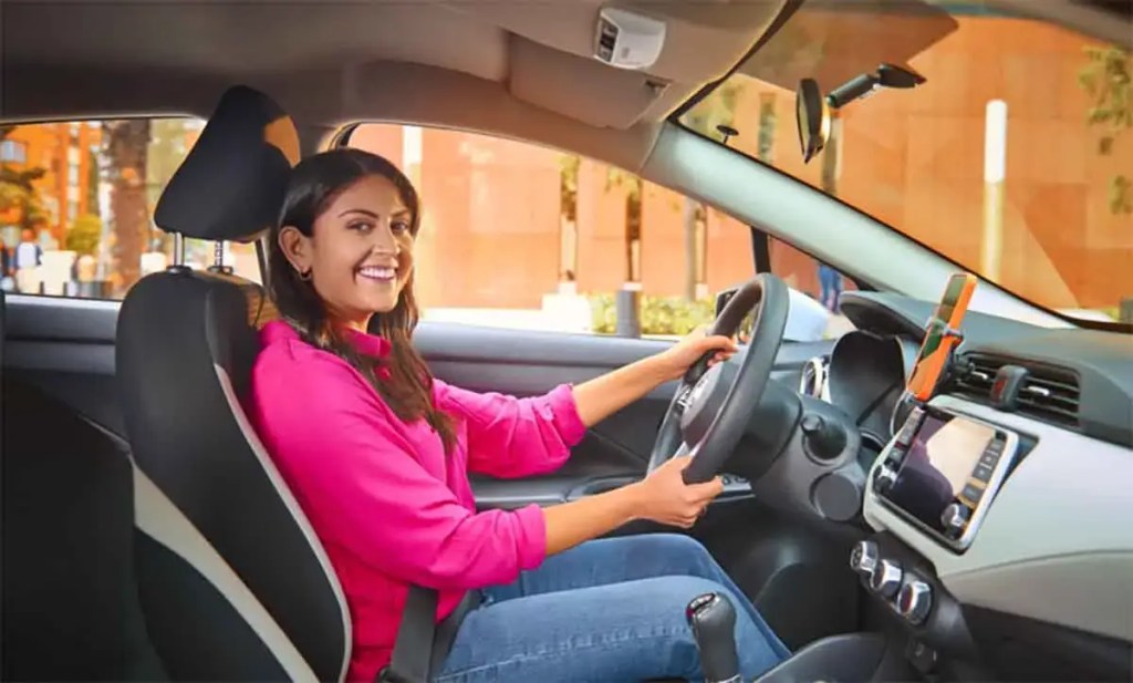 Mujer sonriendo mientras conduce un coche, sentada en el asiento del conductor con un volante en sus manos, vestida con una camisa rosa.