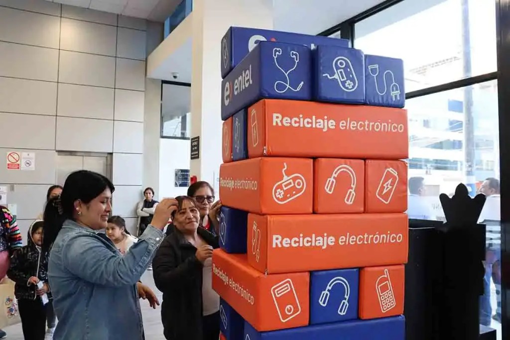 Participantes del concurso de reciclaje del Británico interactuando con un tótem de reciclaje electrónico formado por cajas coloridas, en un ambiente interior.
