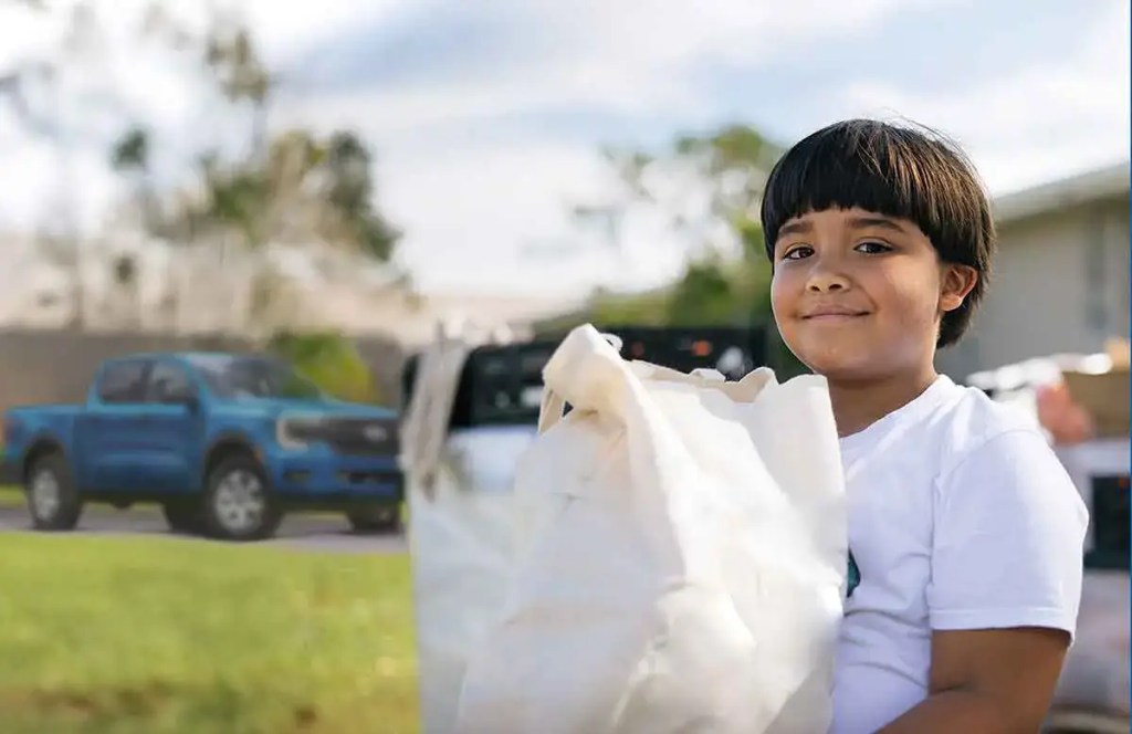 Niño sonriente sosteniendo una bolsa de alimentos, con vehículos Ford en el fondo, en un evento de recolección de alimentos.