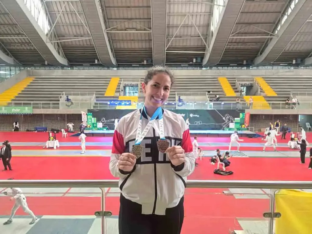 María Luisa Doig, esgrimista peruana, sonríe mientras sostiene sus medallas de oro y bronce en el Campeonato Sudamericano de Esgrima 2025, con una pista de esgrima y competidores al fondo.