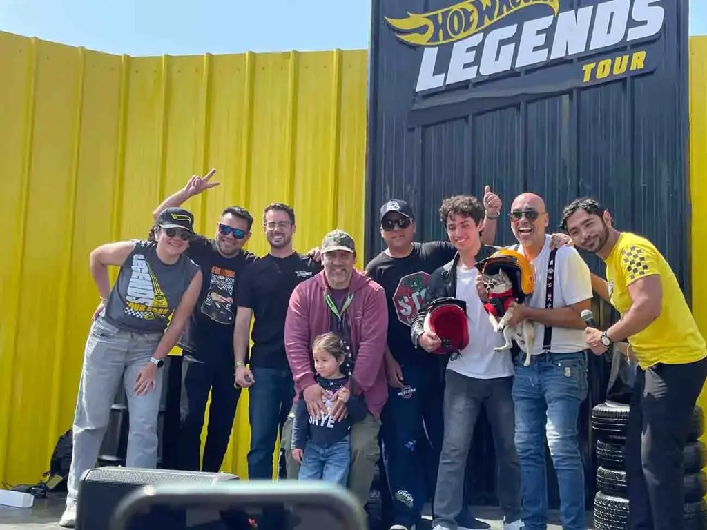 Grupo de personas posando juntas en un evento del Hot Wheels Legends Tour, con un fondo amarillo y el logo del evento visible. Algunas personas sostienen cascos mientras sonríen.