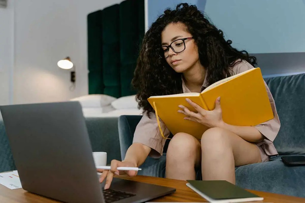 Una estudiante con cabello rizado y gafas, sentada en un sofá, interactuando con una computadora portátil mientras sostiene un cuaderno amarillo.