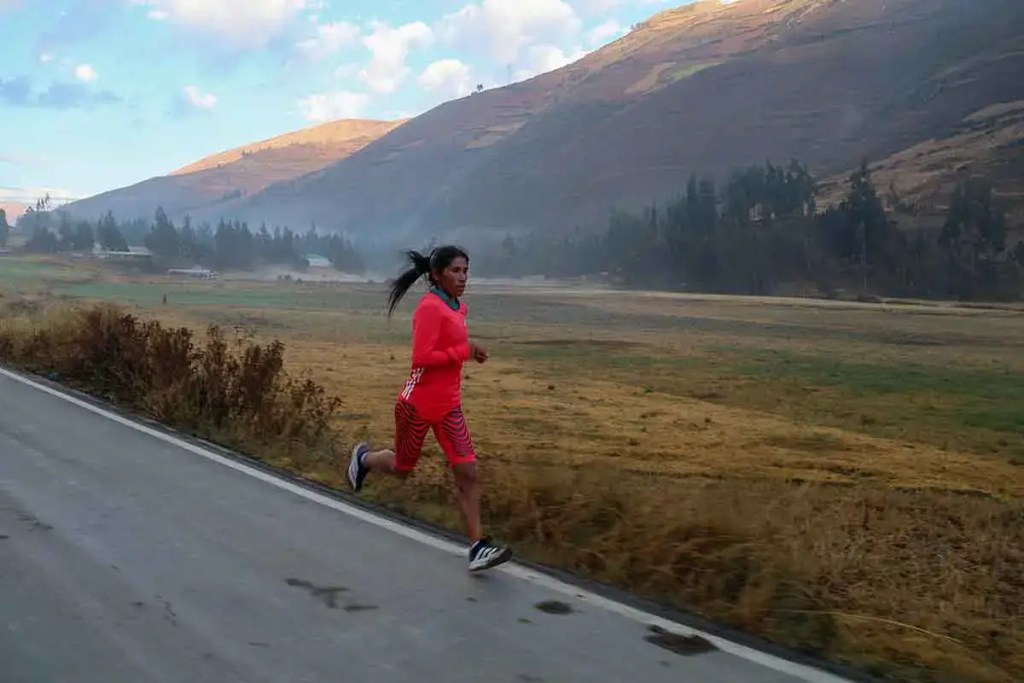 Una maratonista peruana corriendo por una carretera rural en un paisaje montañoso, vistiendo una camiseta y pantalones cortos rojos.
