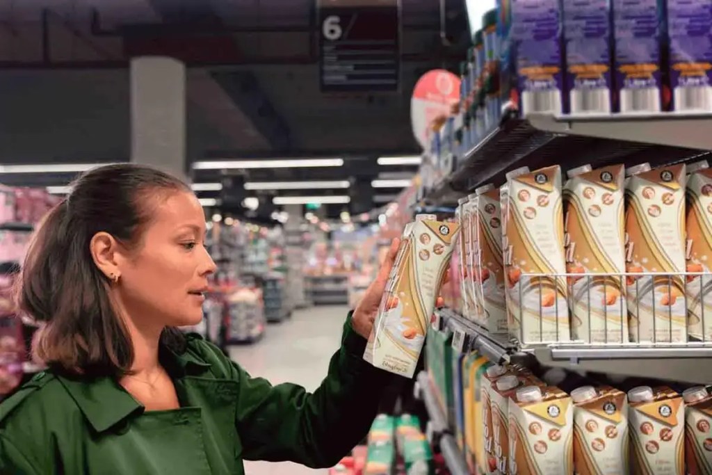 Una mujer observa un envase de cartón en un estante de supermercado lleno de productos.