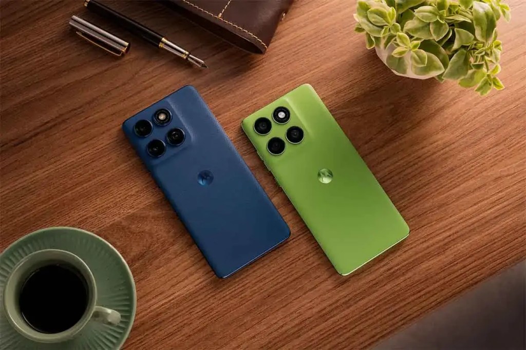 Dos smartphones Motorola de colores azul y verde sobre una mesa de madera, junto a una taza de café y una planta pequeña.