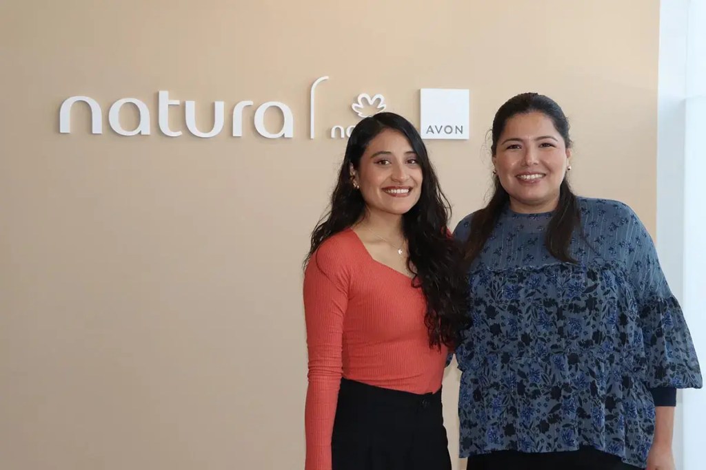 Dos consultoras de belleza sonrientes posan frente al logotipo de Natura y Avon en una pared beige.
